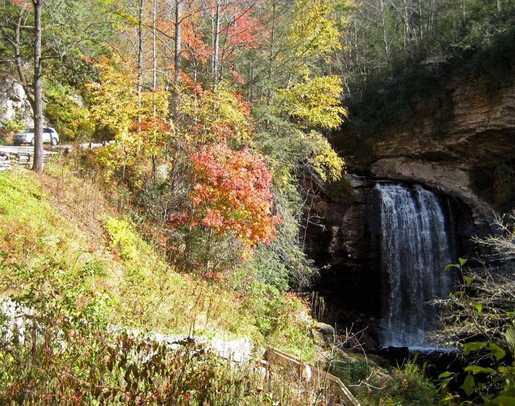 Amidst colorful autumn leaves, a vehicle travels along the Forest Heritage Scenic Byway next to Looking Glass Falls, a 30-foot wide, 60-foot high waterfall. The route of the byway was originally a logging railroad route used to transport timber to a nearby sawmill.