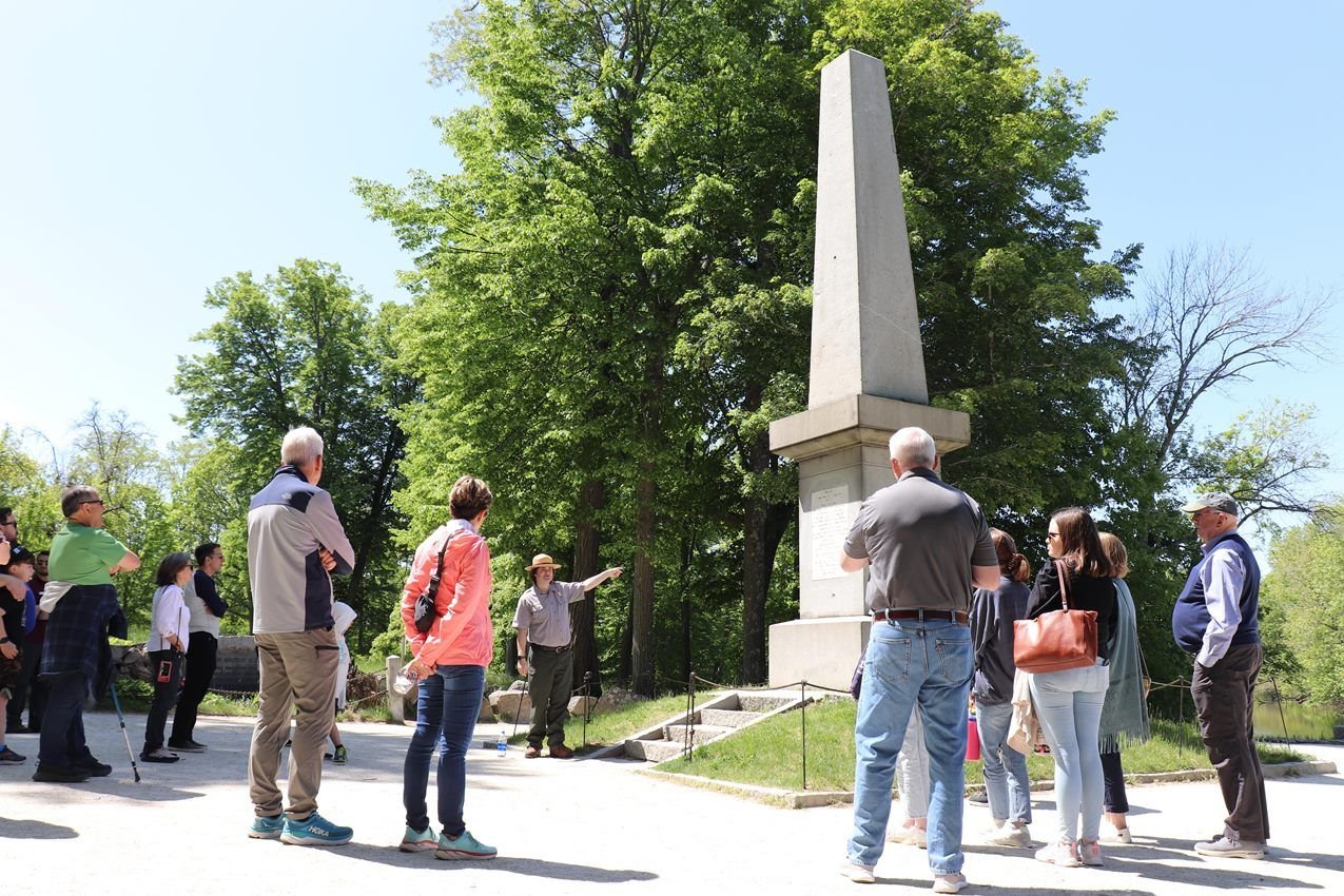 A park ranger stands in front of a stone obelisk monument pointing and talking to visitors