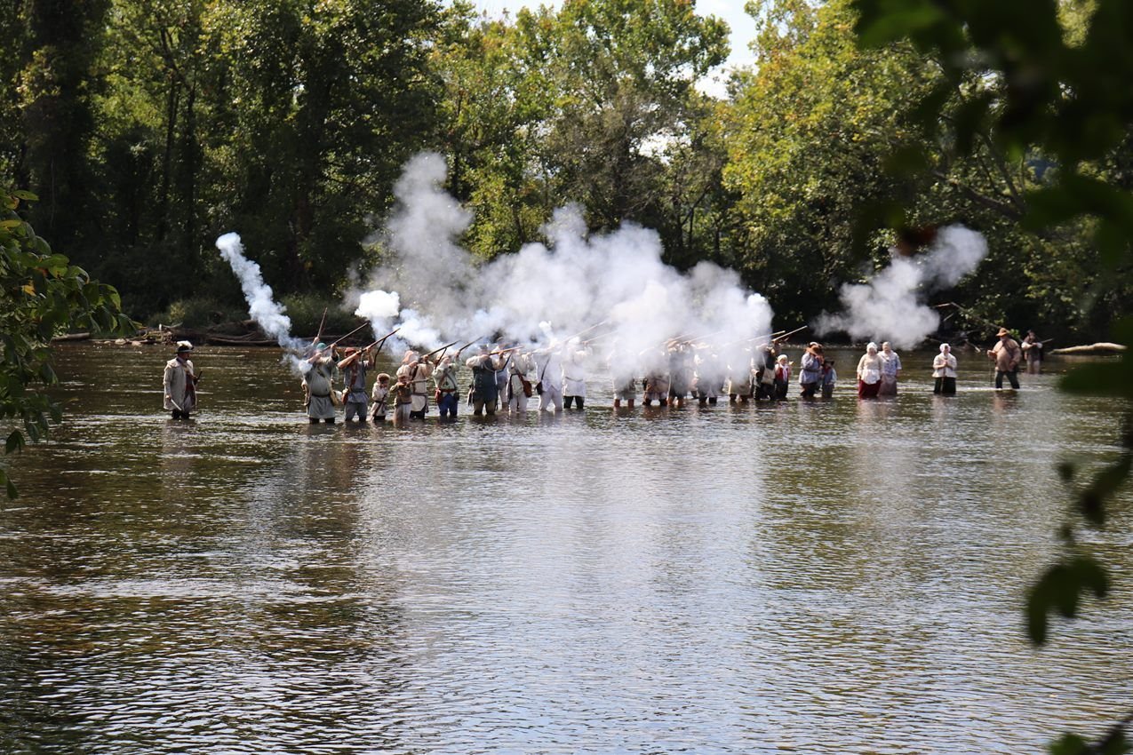 A large group of living historians stand in a river and fire muskets, with a cloud of smoke rising.