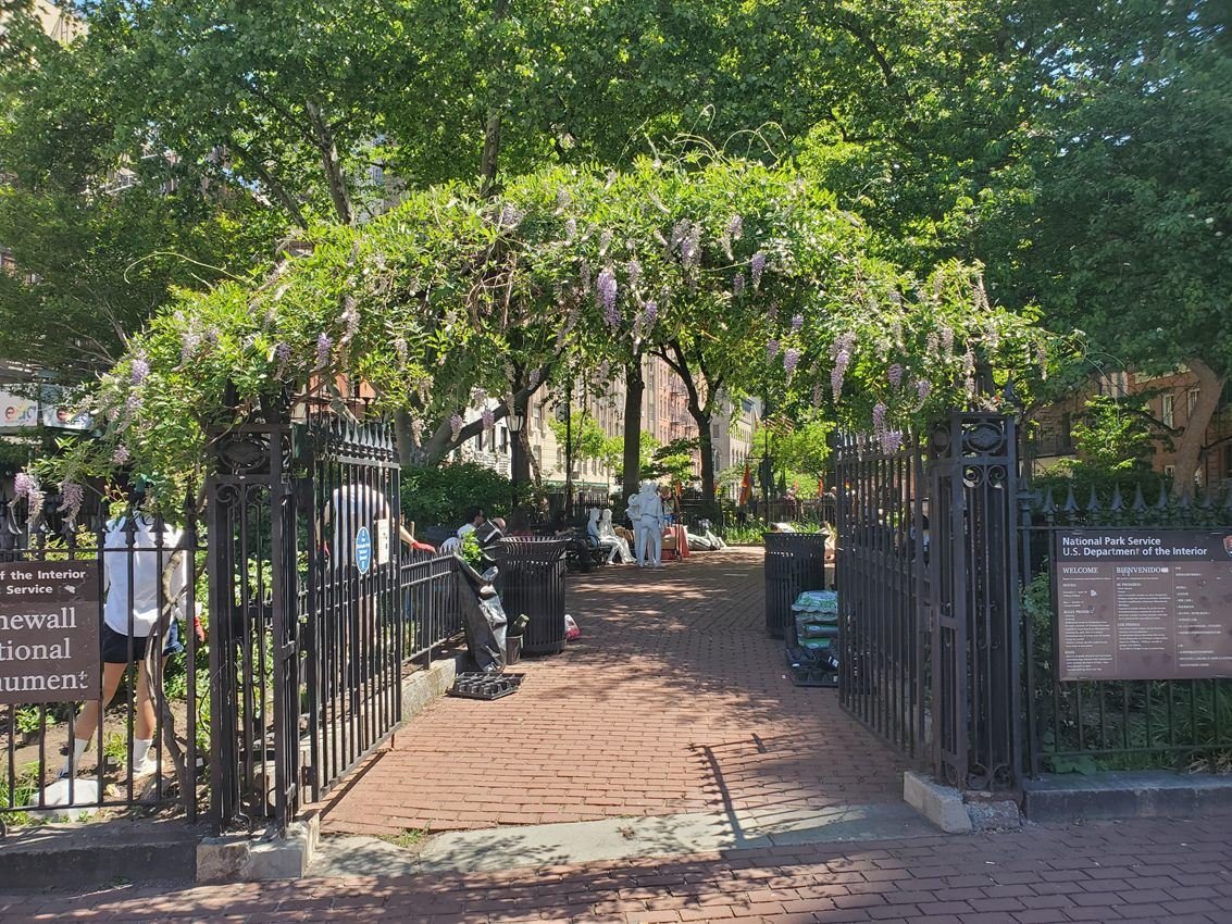 An open black fence gate with green plants blooming above it.