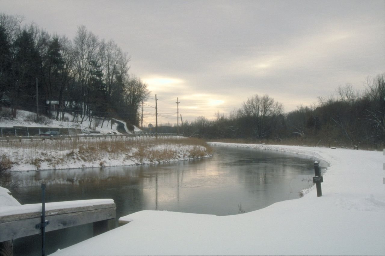 A canal behind the Canal Visitor Center in Cuyahoga Valley National Park is partially iced over during the winter. There is snow on the ground, and the remote sun is setting.