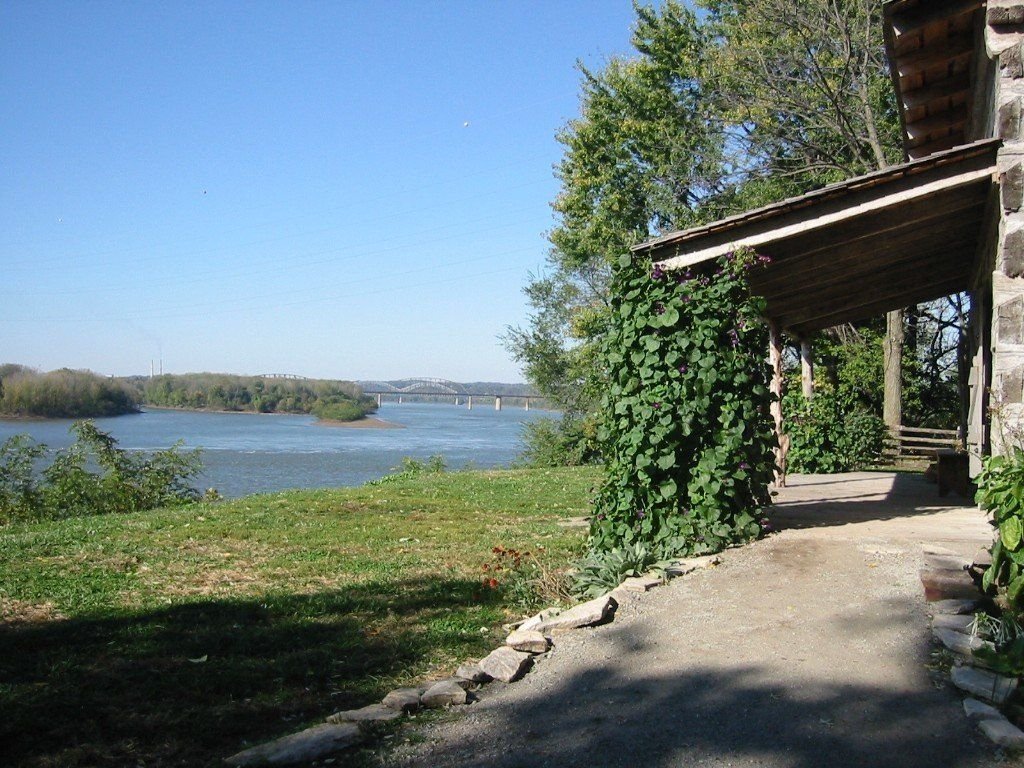 The waters of the Ohio River run blue, as seen from the vine-covered porch of the Clark replica cabin; in the distance the Louisville/New Albany bridge spans the river.