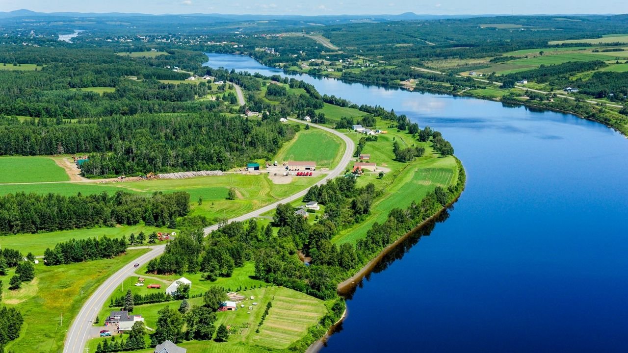 Far reaching aerial view of the deep blue highway and undulating St. John River – the international boundary – looking westward on a beautiful summer day. US Route 1 hugs the river’s south shore winding by scattered buildings and a patchwork of greens.